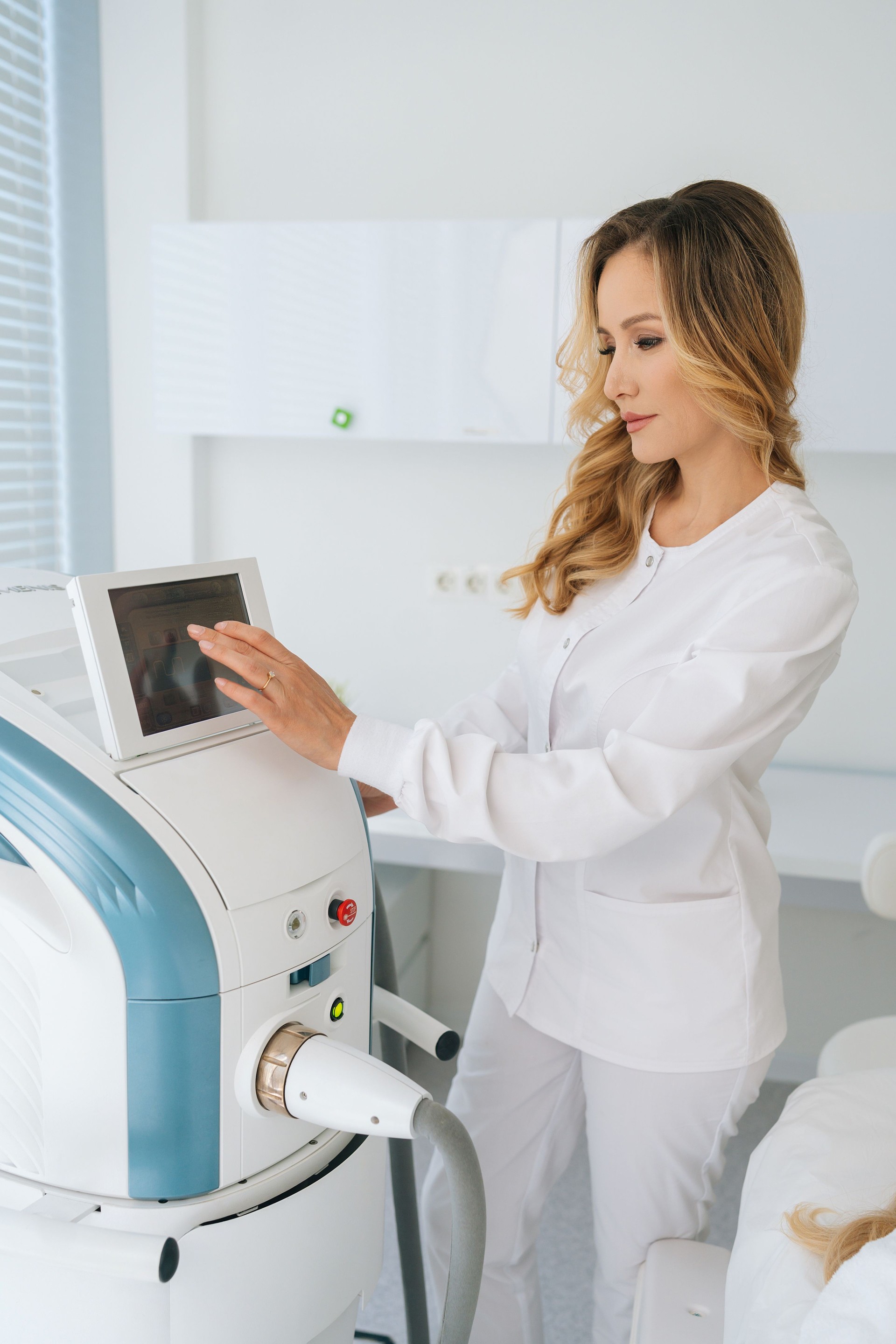 Vertical shot of professional female cosmetologist adjusts device for photorejuvenation using touchscreen in beauty clinic.